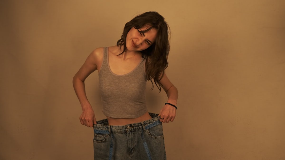 Smiling young woman in oversized jeans celebrates weight loss accomplishment.